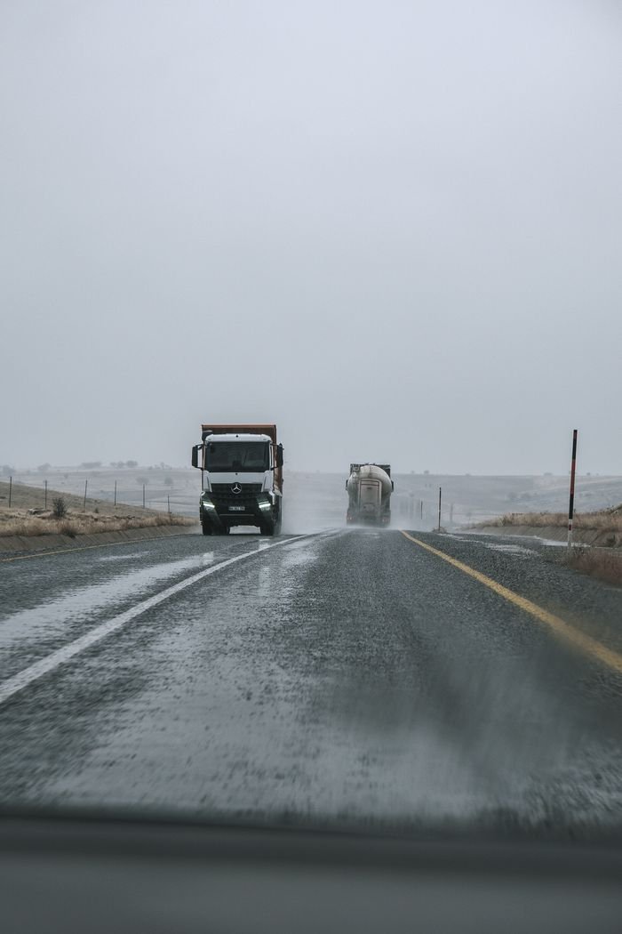 Two trucks driving on a rainy highway with overcast skies and empty roadsides.