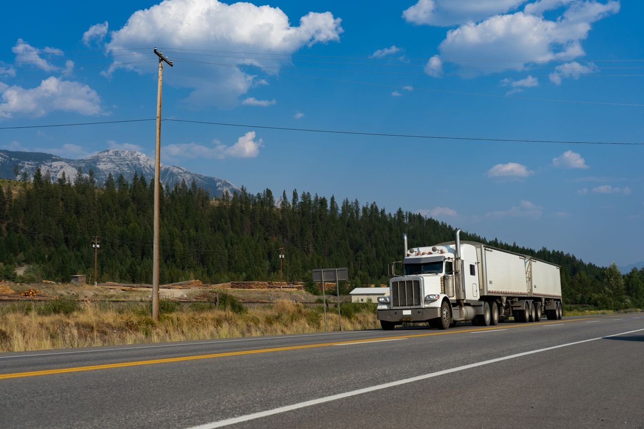 A white semi-truck traveling on a scenic road in Jaffray, BC, with mountains in the background.